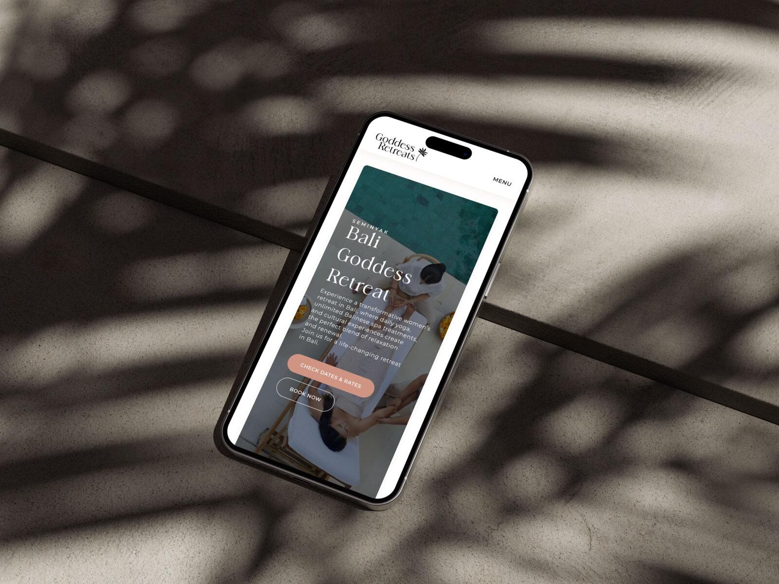 A mobile phone on concrete displaying the retreat booking page design for the Goddess Retreats website.