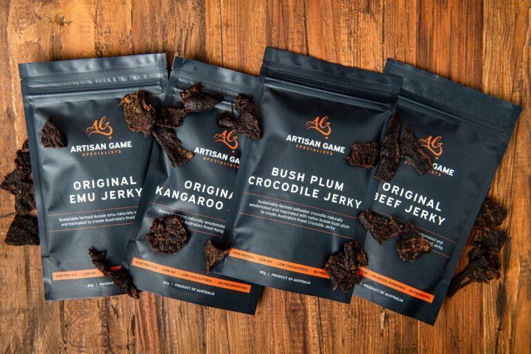 Four bags of Artisan Game jerky packaging are arranged on a table, with pieces of jerky scattered around them.