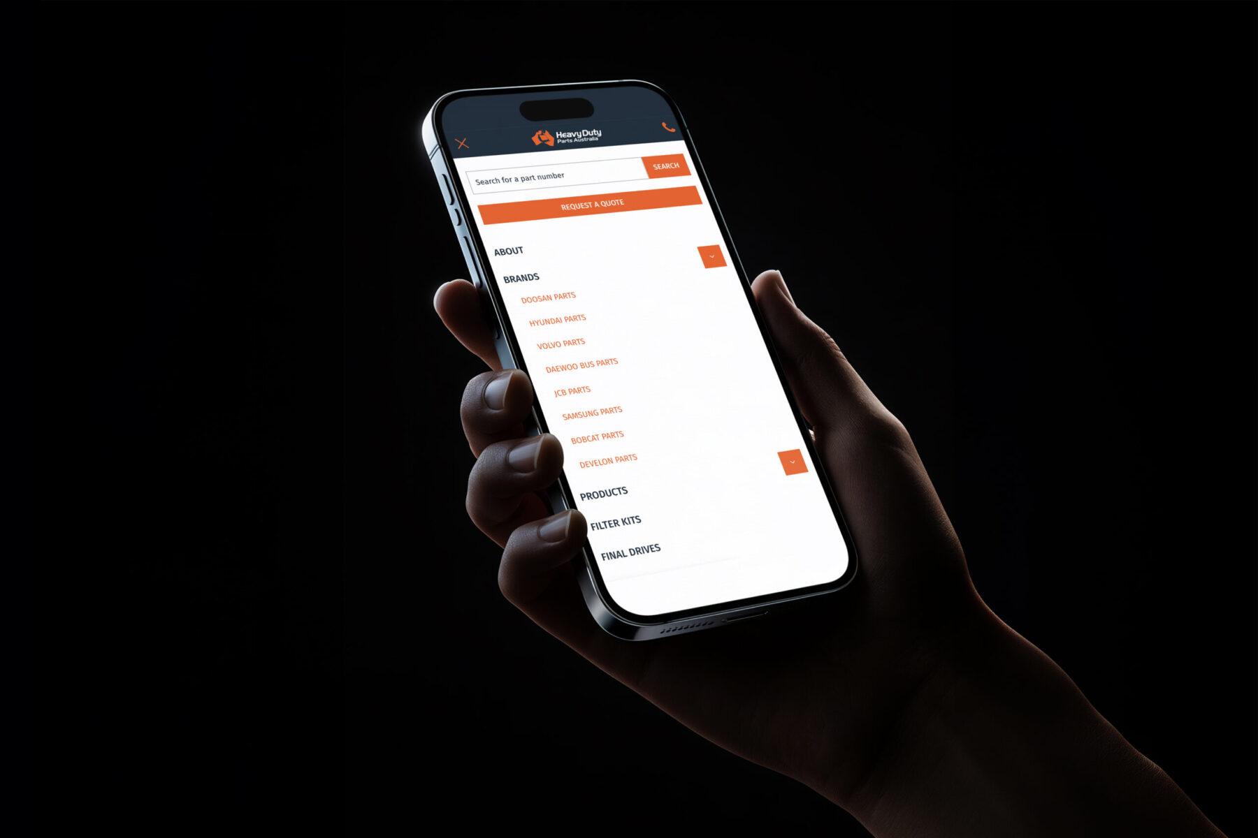 A mockup of a mobile phone being held up in a dark room, displaying the Heavy Duty Parts Australia website navigation design.